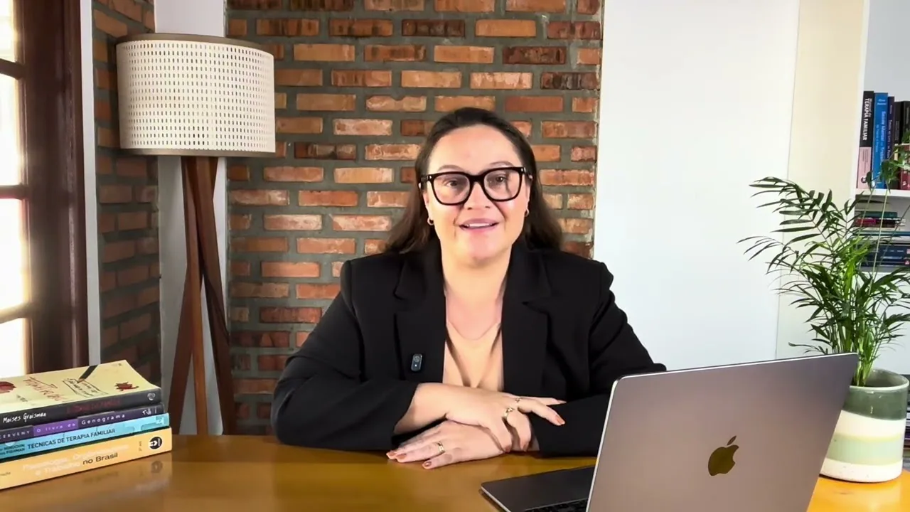 Dra. Karine Schwaab sentada a uma mesa de madeira, de blazer preto e óculos, sorrindo para a câmera; à esquerda, uma pilha de livros, e à direita um notebook aberto, diante de uma parede de tijolos com uma luminária de piso ao lado.