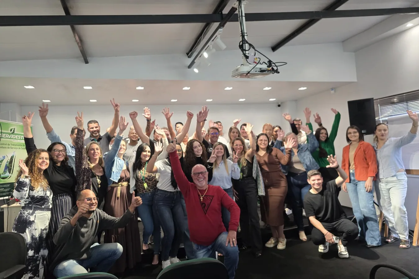 Grupo de cerca de trinta participantes com os braços erguidos, em uma sala de treinamento com um banner corporativo ao fundo.