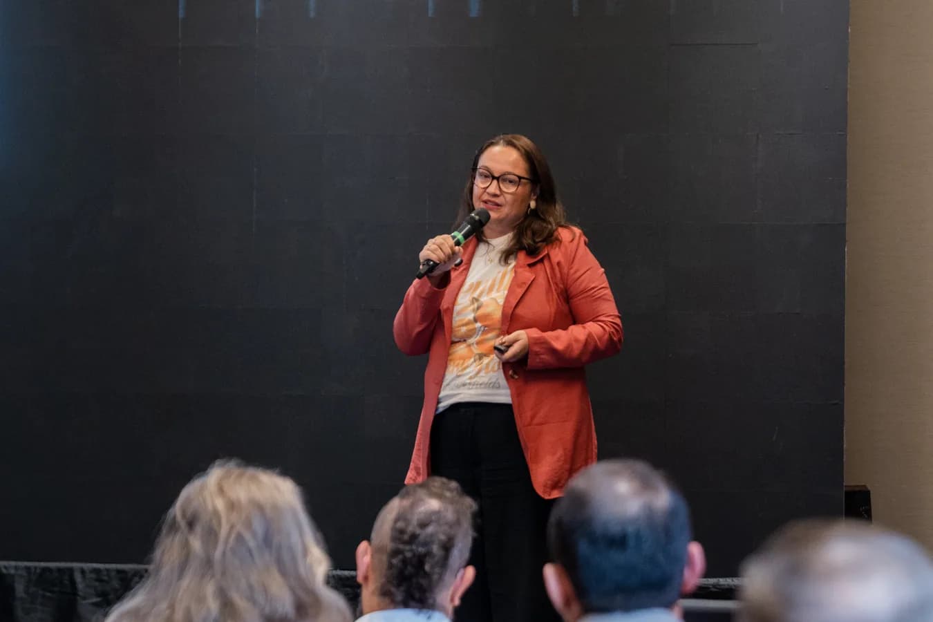 Karine Schwaab ao microfone, em pé diante de um painel escuro; três pessoas da plateia aparecem de costas em primeiro plano.