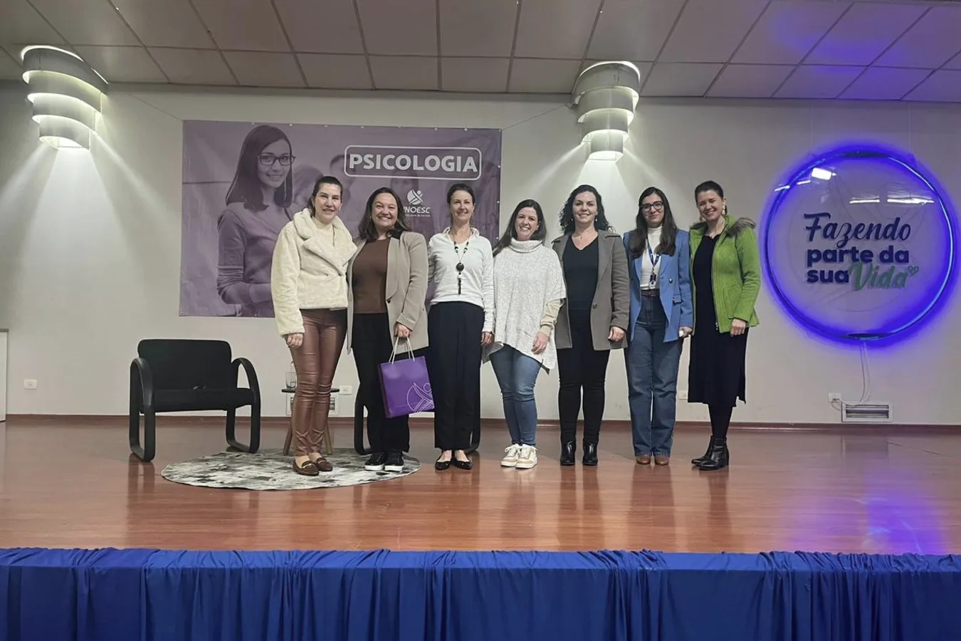 Sete mulheres em pé no palco de um auditório, diante de um banner de um curso de Psicologia.